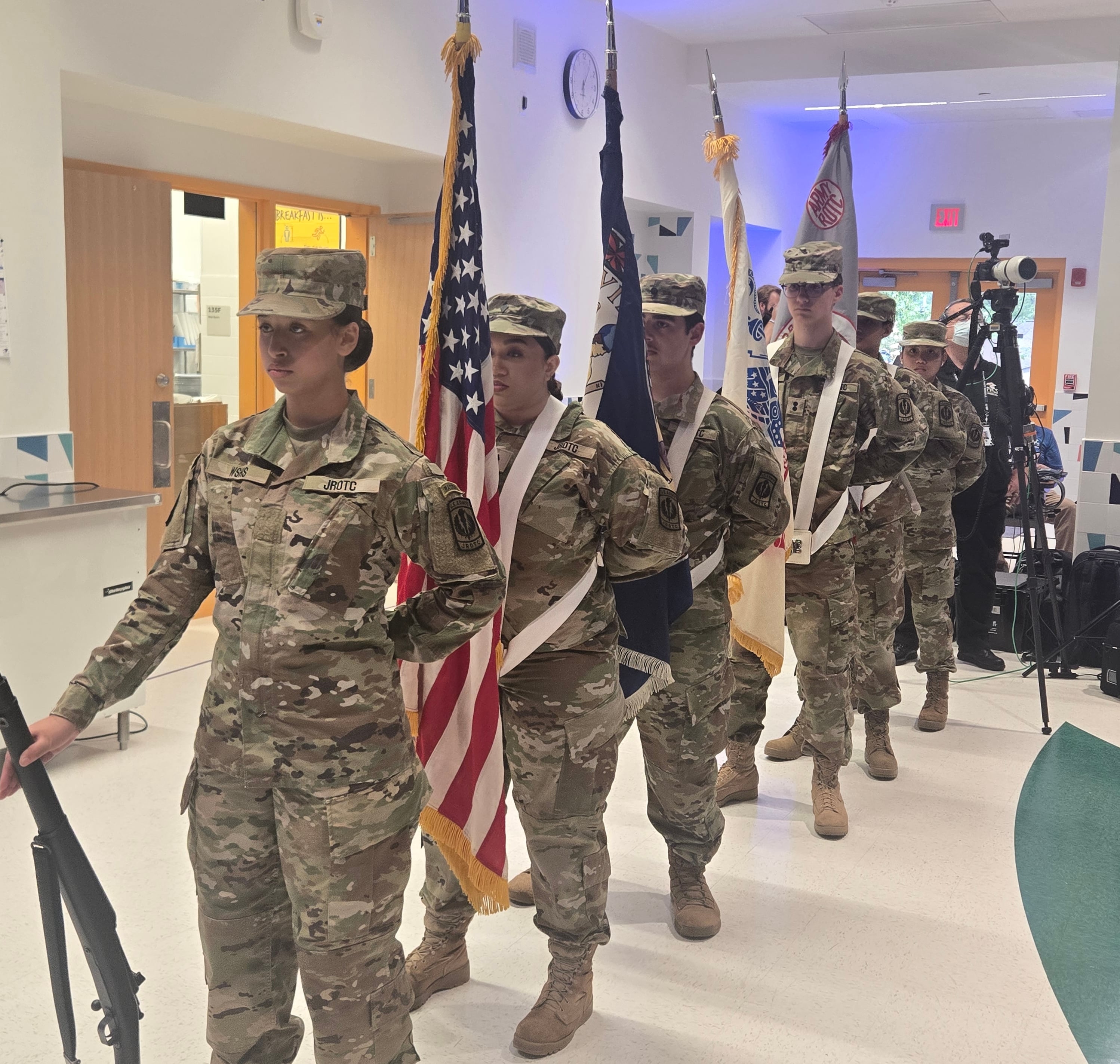 JROTC color guard in camouflage uniforms marches indoors carrying the U.S. flag and other ceremonial flags.