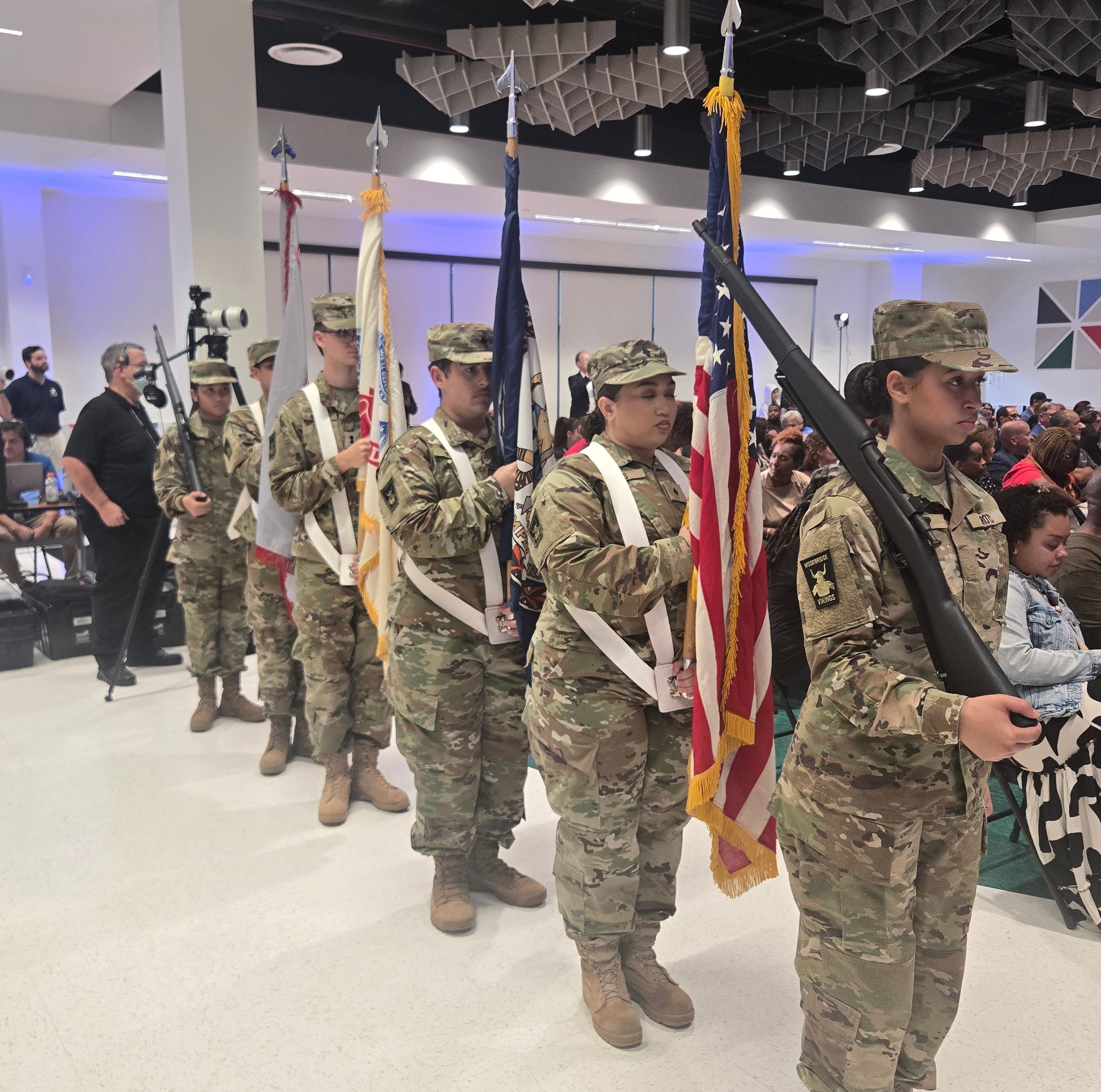 Military color guard in camouflage uniforms marches indoors carrying the U.S. flag and other ceremonial flags as a seated audience watches.