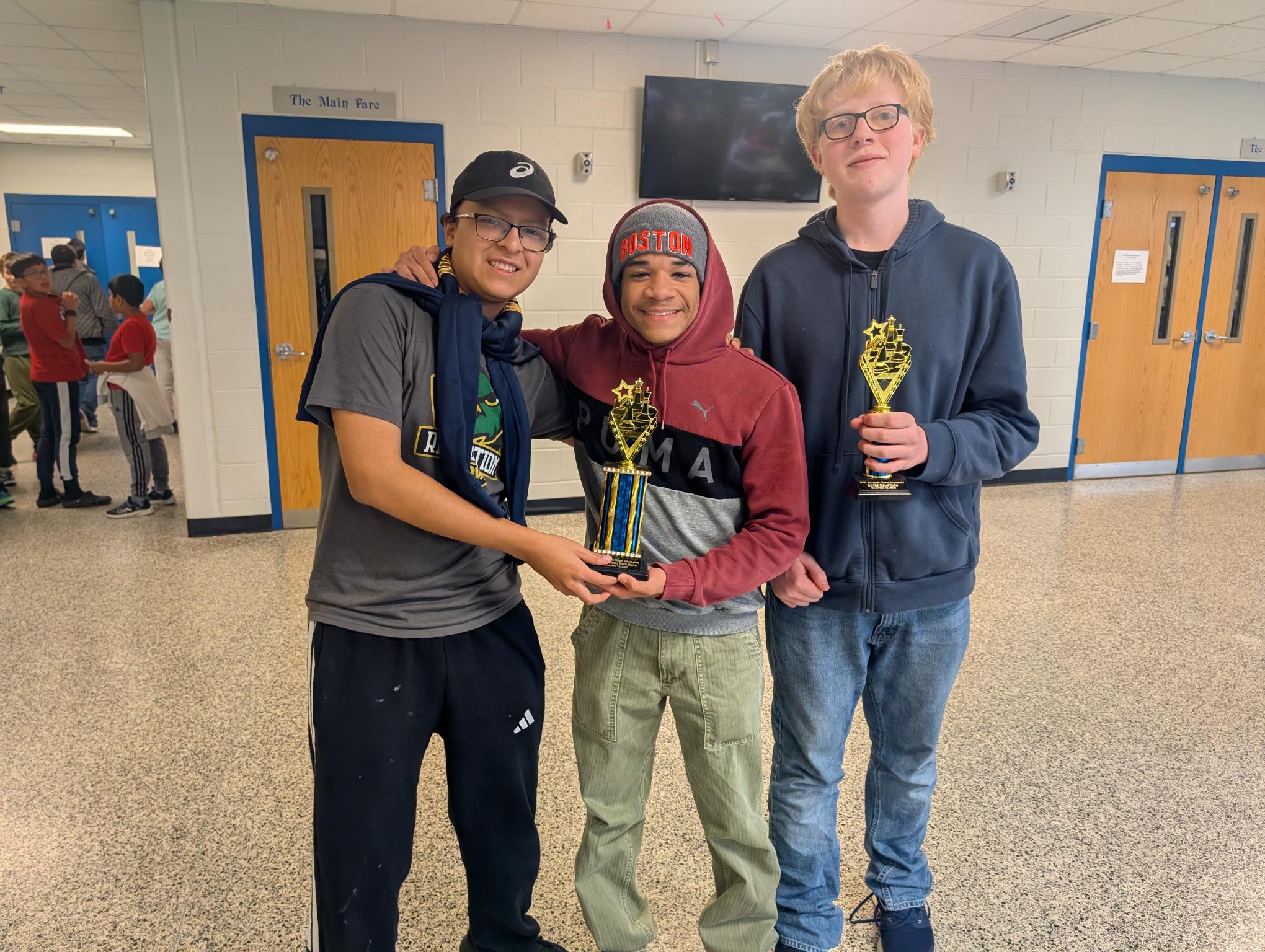 Three students proudly hold their trophies in a school hallway, celebrating their achievements.