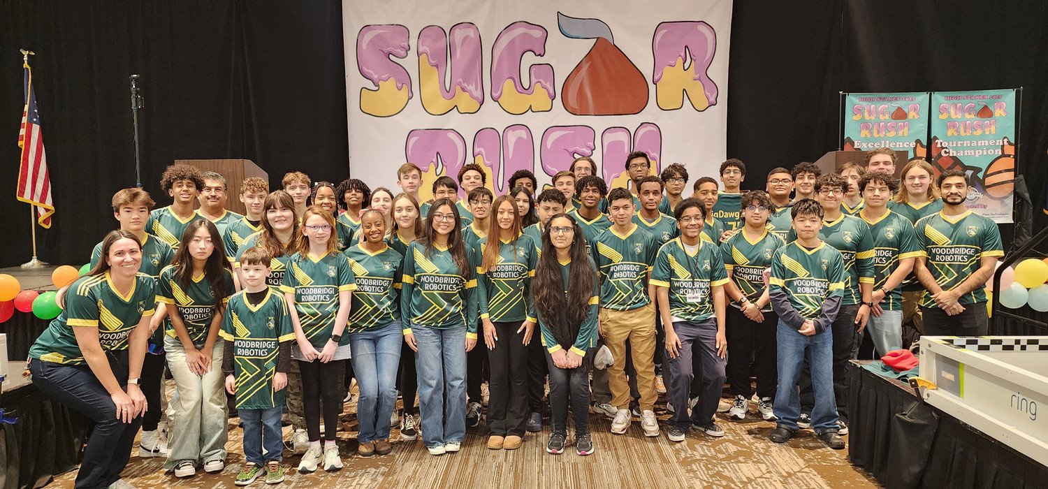 The WSHS Robotics Team wearing matching green team shirts poses proudly in front of a colorful "Sugar Rush" backdrop at a robotics tournament.
