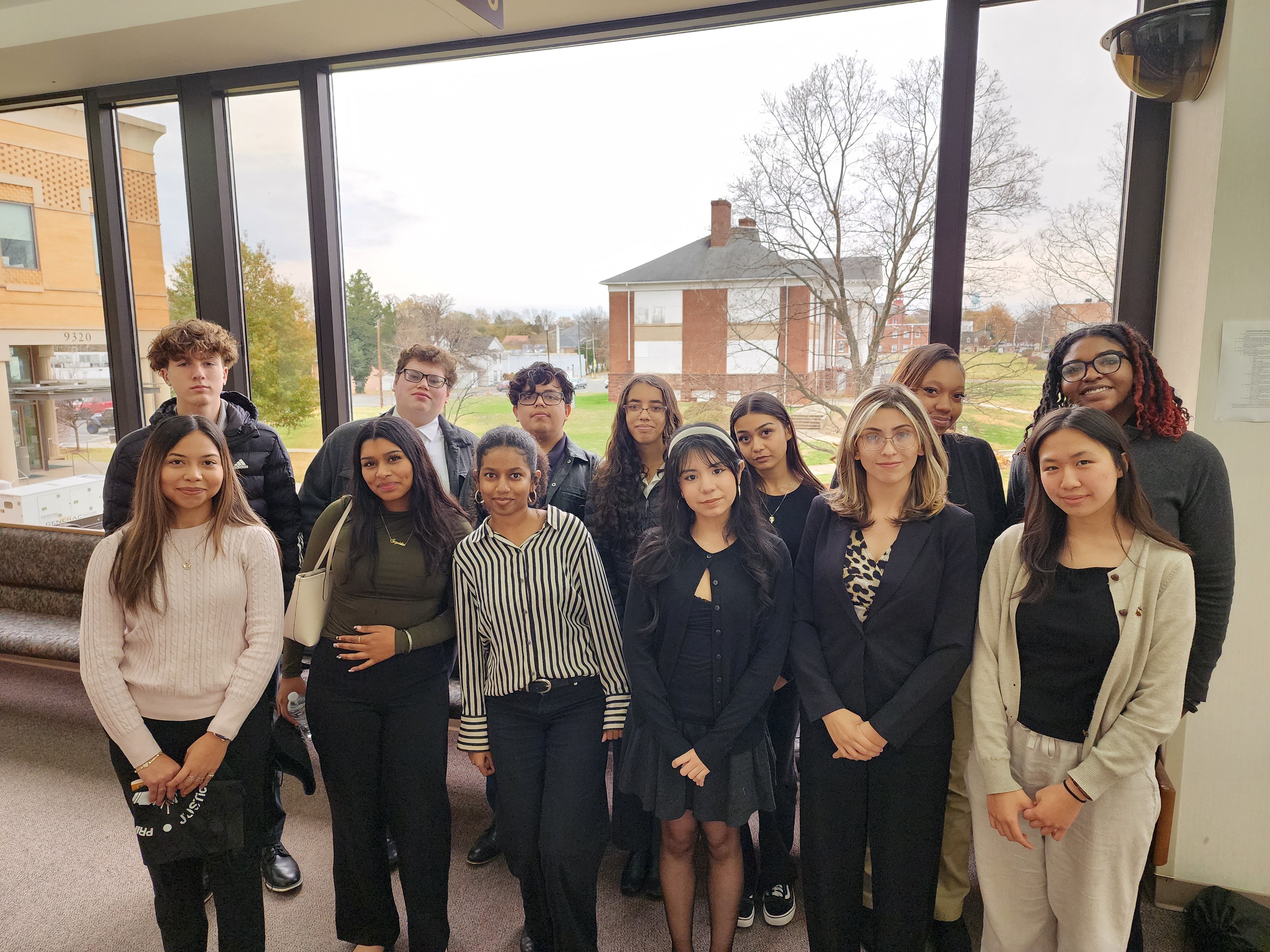 Group photo of students dressed in formal attire, posing indoors with a view of an outdoor pavilion in the background.