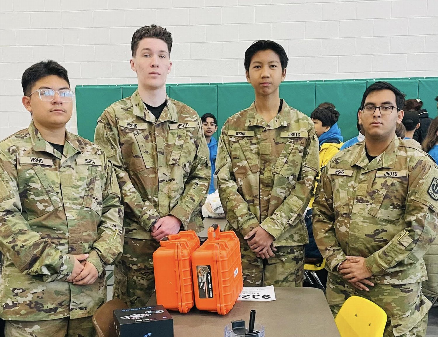 Four cadets in military camouflage uniforms pose at a table with orange equipment cases and other items.