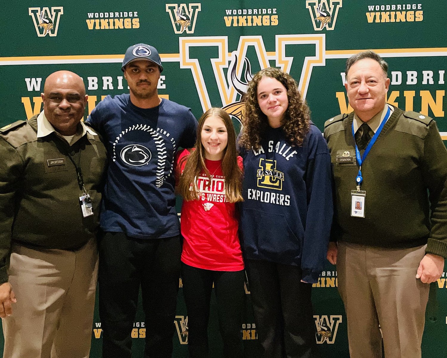 A group photo of four individuals posing together, including students wearing school spirit shirts and two staff members in uniforms, in front of a Woodbridge Vikings backdrop.