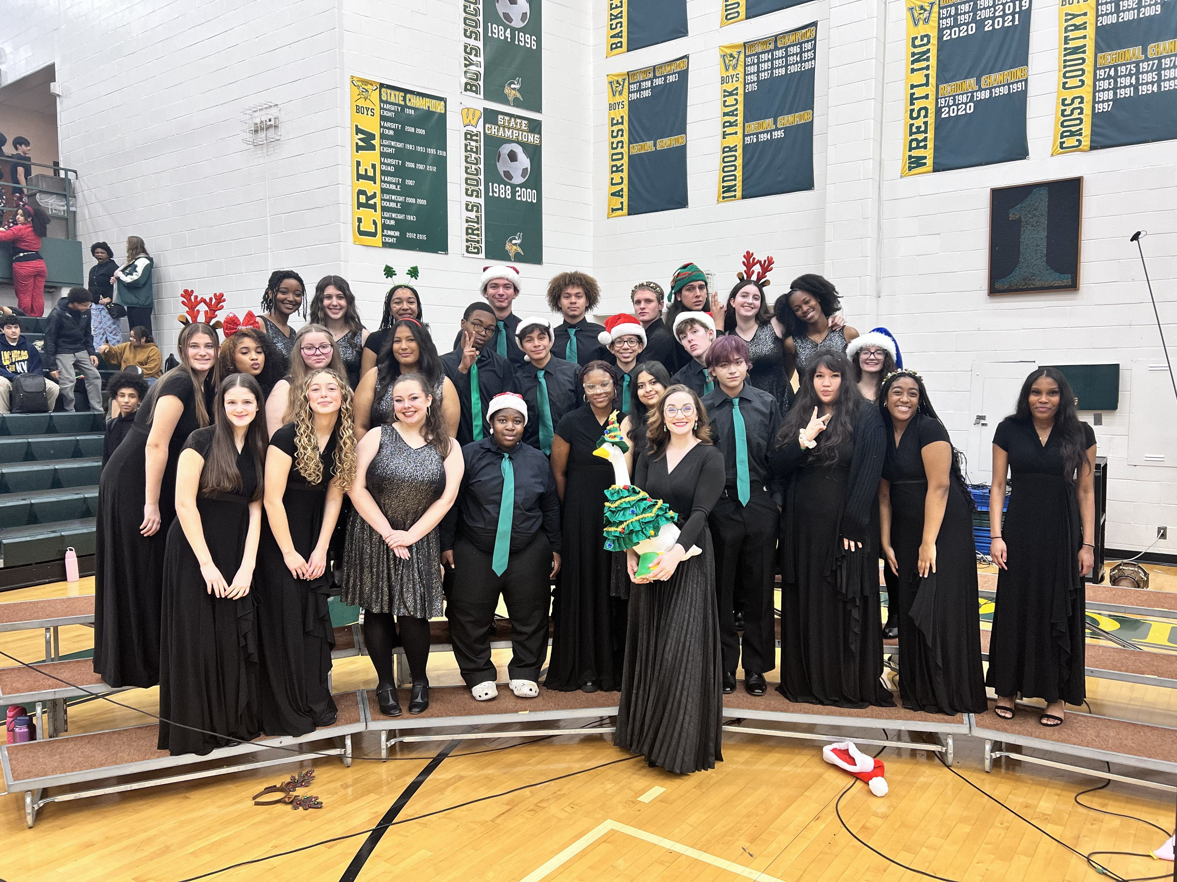 A group photo of a choir dressed in formal black attire, posing together with holiday-themed accessories and decorations in a school gymnasium.