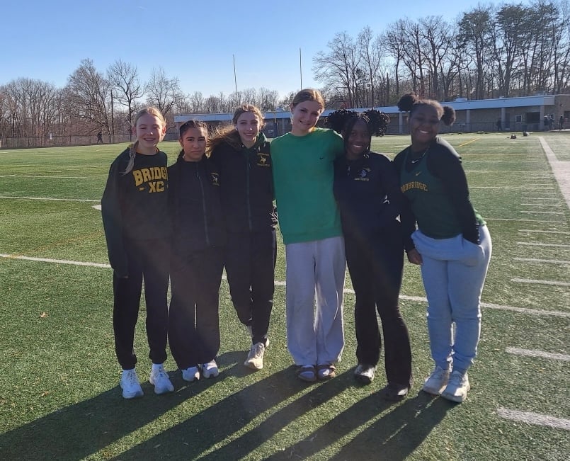 A group of six female athletes stands together on a sports field, wearing team apparel and smiling for the camera.