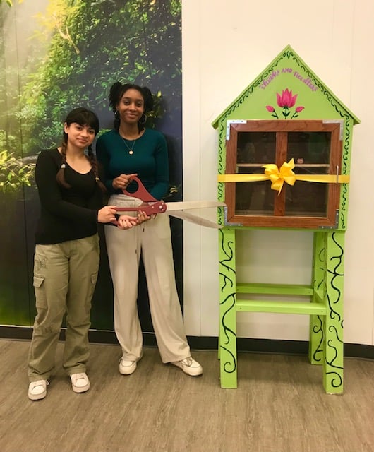 Naima and Edria stand next to a brightly painted green little free library, one holding large scissors poised to cut a yellow ribbon.
