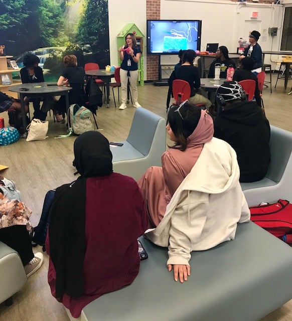 A group of students attentively listens to a crocheting presentation in a lively learning environment.