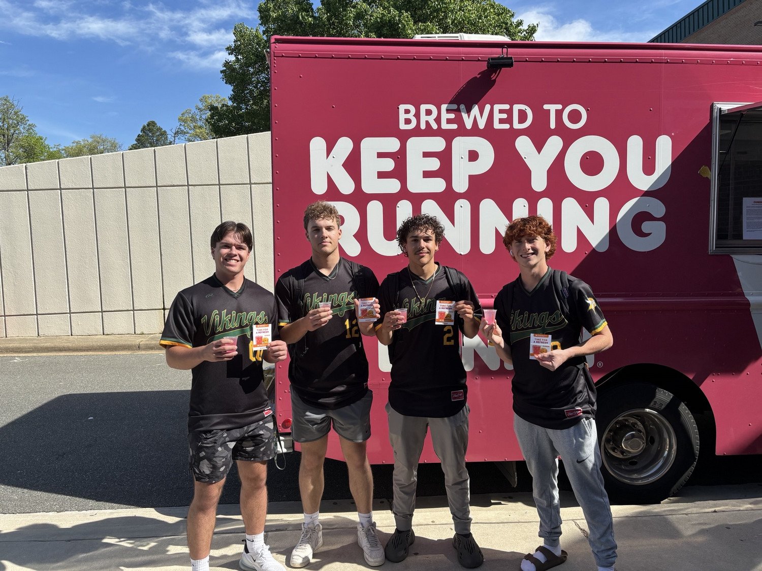 Four young men in team jerseys pose in front of a food truck, all holding cups of juice. The truck has a pink exterior with the slogan "Brewed to Keep You Running."
