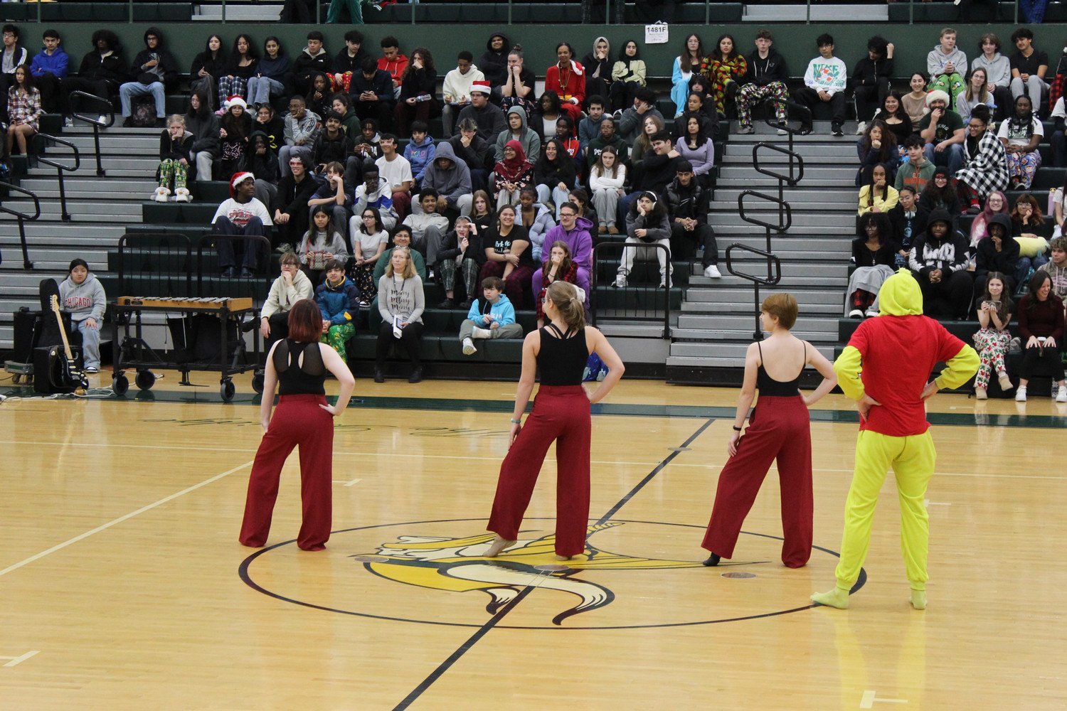 A dance performance featuring four dancers in colorful costumes on a gymnasium court, with an audience seated in the bleachers watching intently.