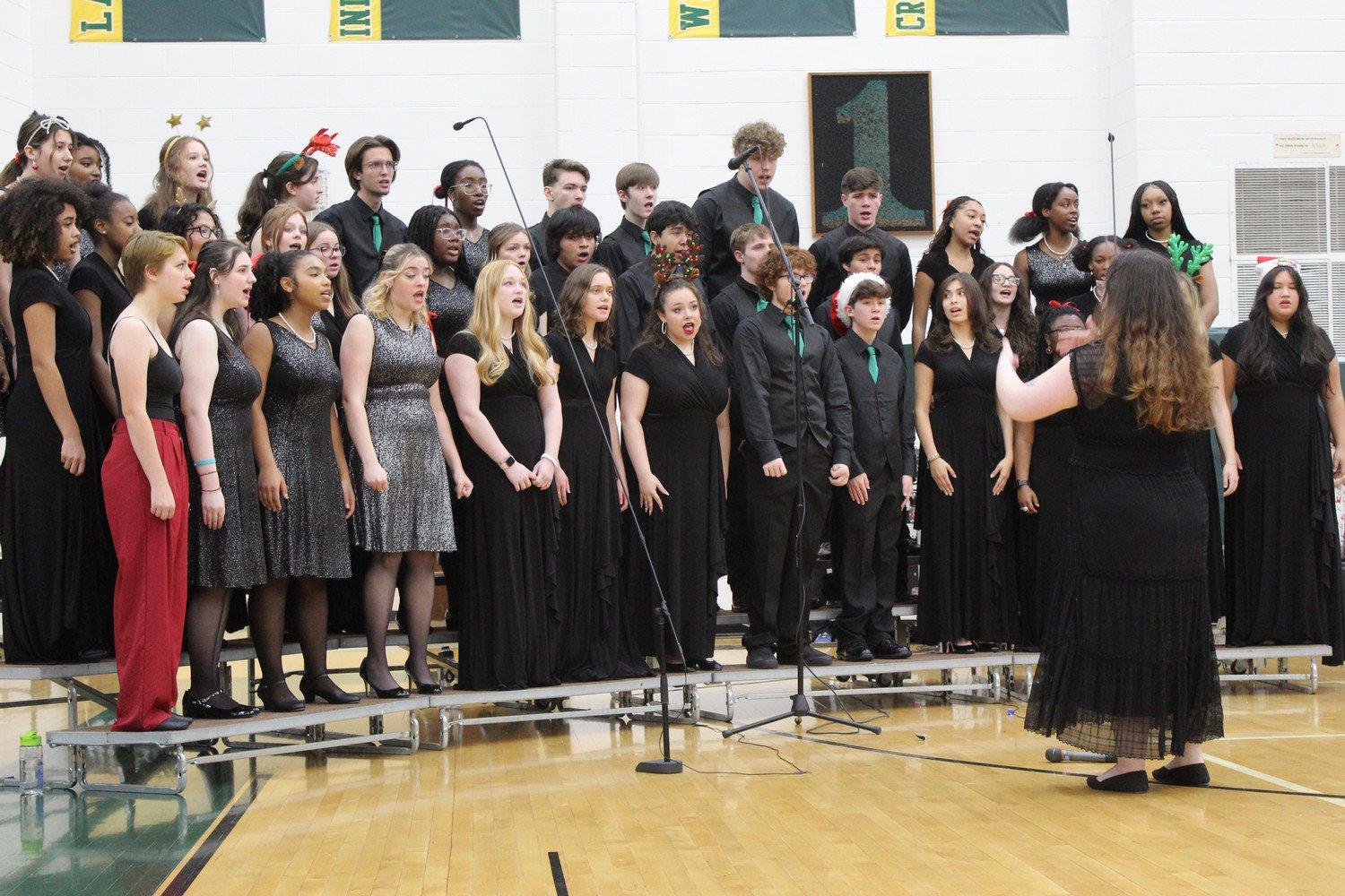 A high school choir performing onstage, dressed in black attire, with a conductor leading them.