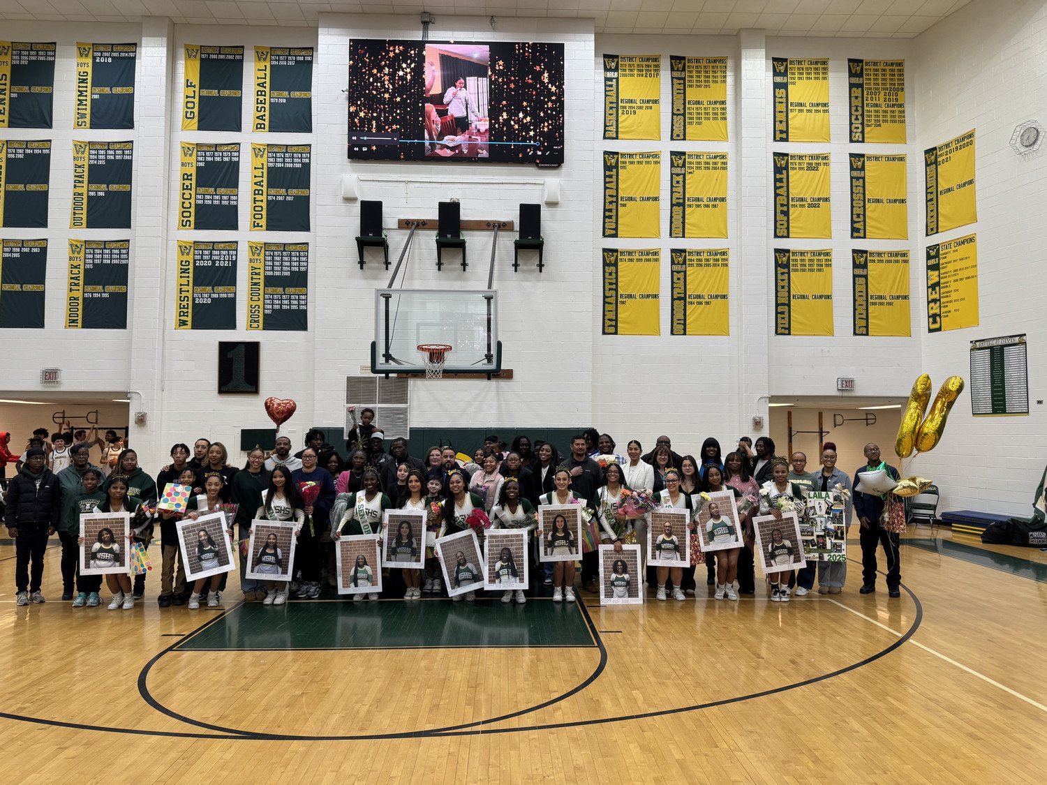 WSHS Cheerleaders gathered on a gymnasium floor, holding framed photos and celebrating a special event, with a scoreboard displaying a colorful image above them.