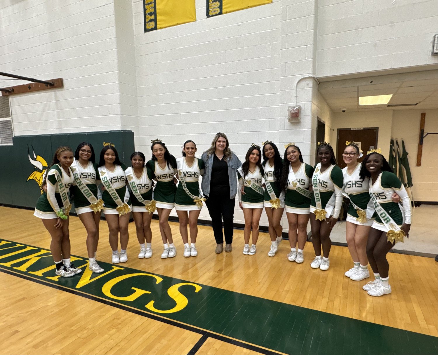WSHS cheerleaders in green and gold uniforms poses together on a gymnasium floor, alongside a woman in a gray jacket.