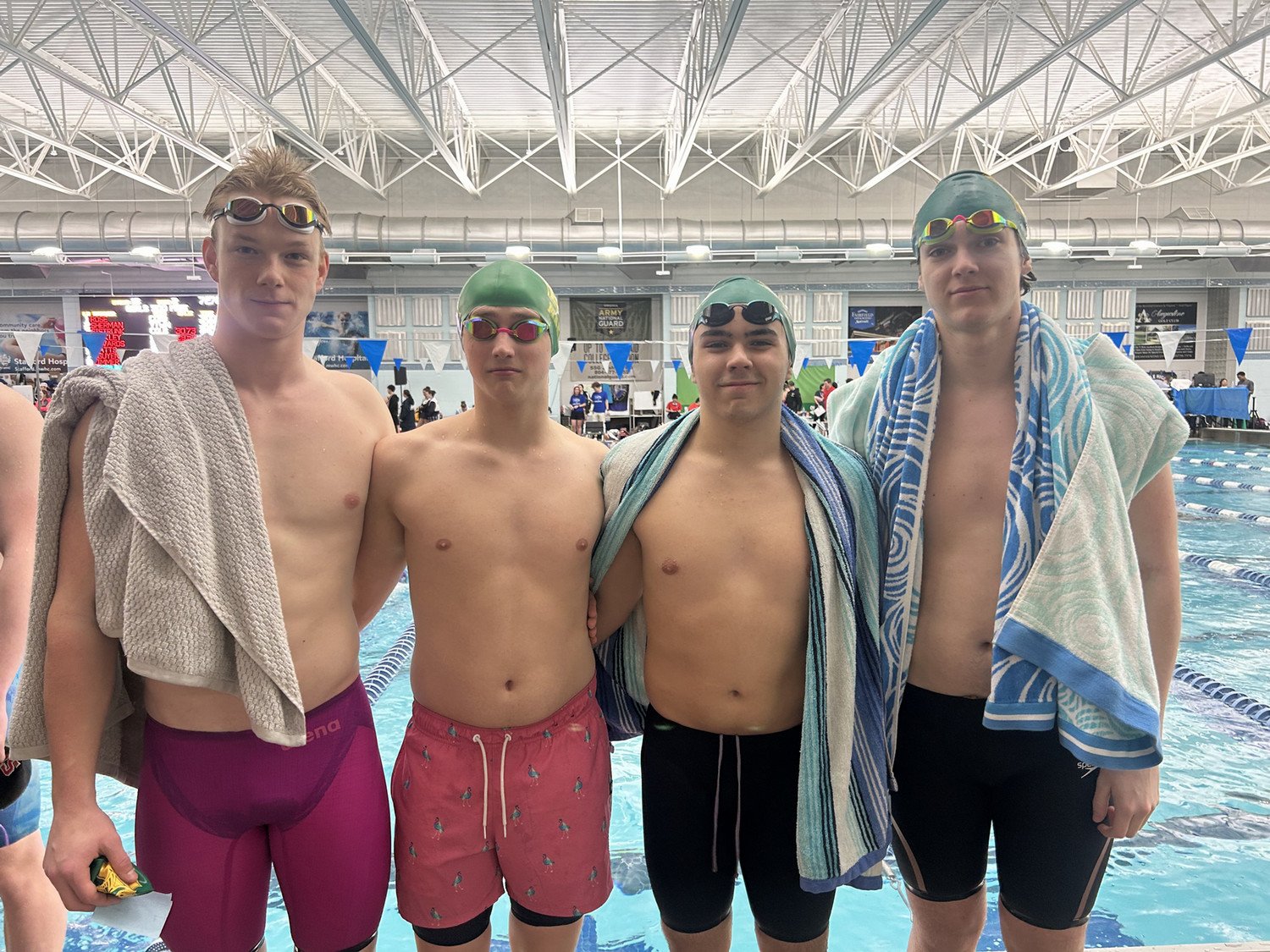 WSHS male swimmers pose together at a swimming competition, wearing swim caps and goggles, with towels draped over their shoulders.
