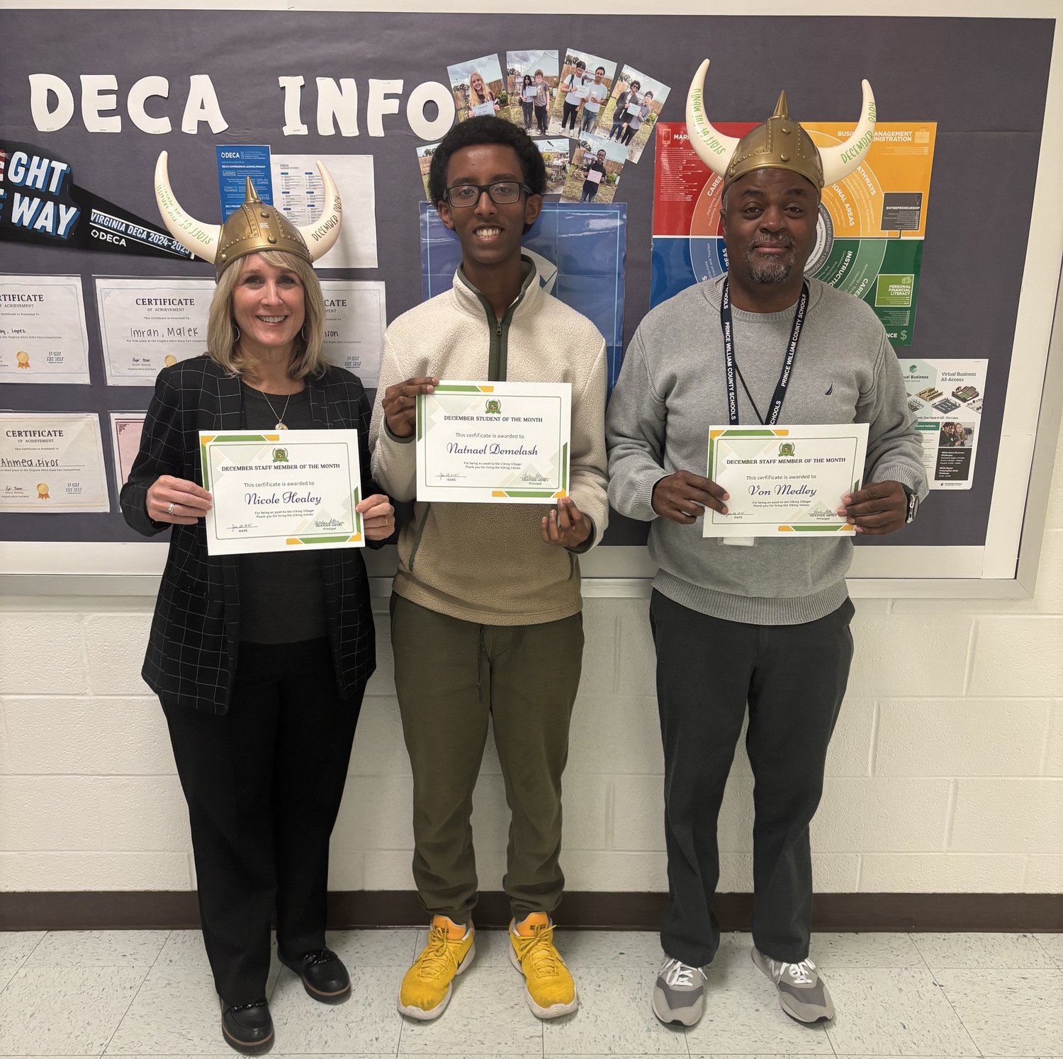 December staff and students of the month, Nicole Healey, Von Medley and Nate Demalash, wearing Viking helmets proudly hold certificates in front of a bulletin board displaying various achievements and information.