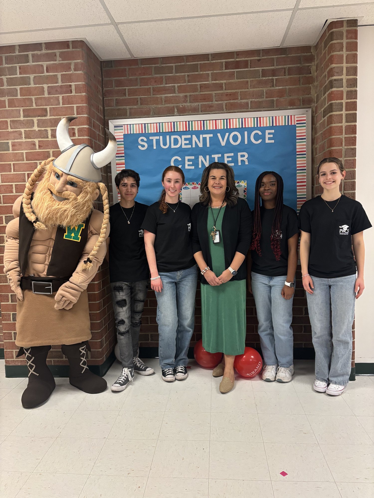 A group of five individuals, including a person in a viking mascot costume, stand in front of a colorful banner for the "Student Voice Center."