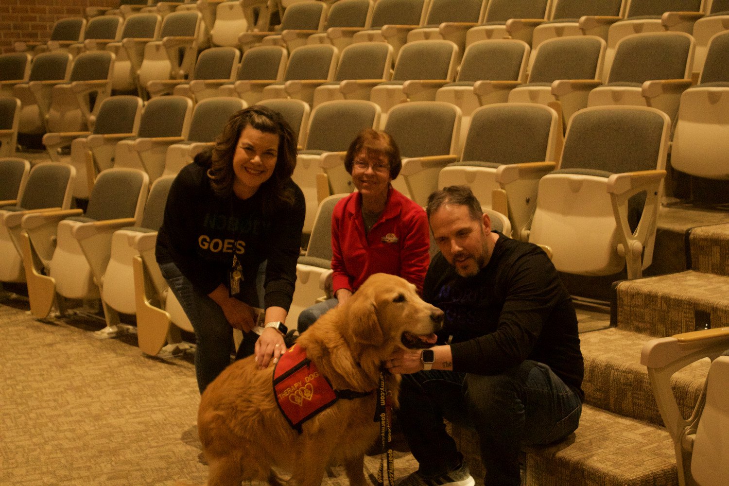 A golden retriever in a service vest sits with three people in an auditorium setting.