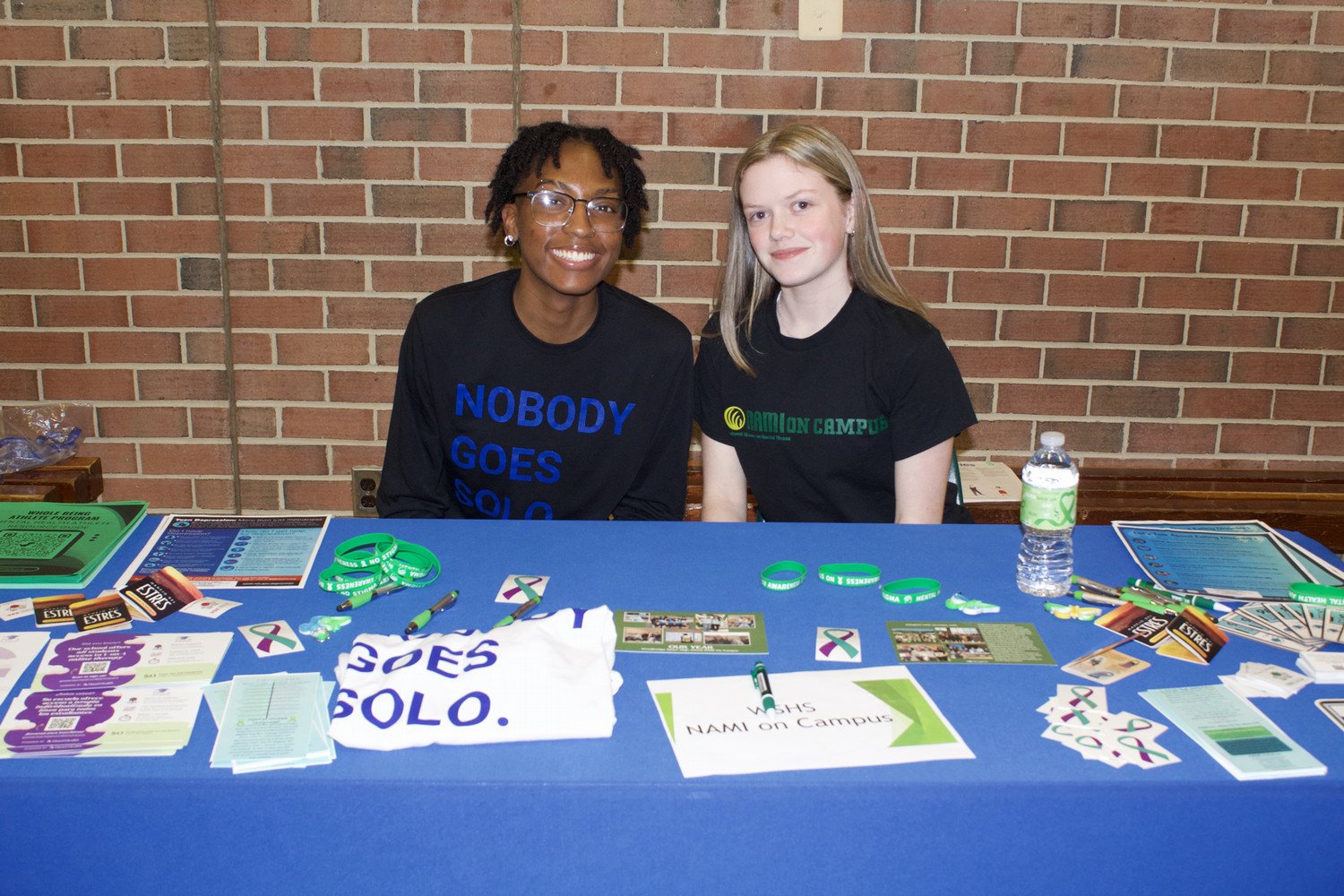 Two students sitting behind a table displaying information and promotional materials related to NAMI on Campus, wearing shirts that read "NOBODY GOES SOLO."