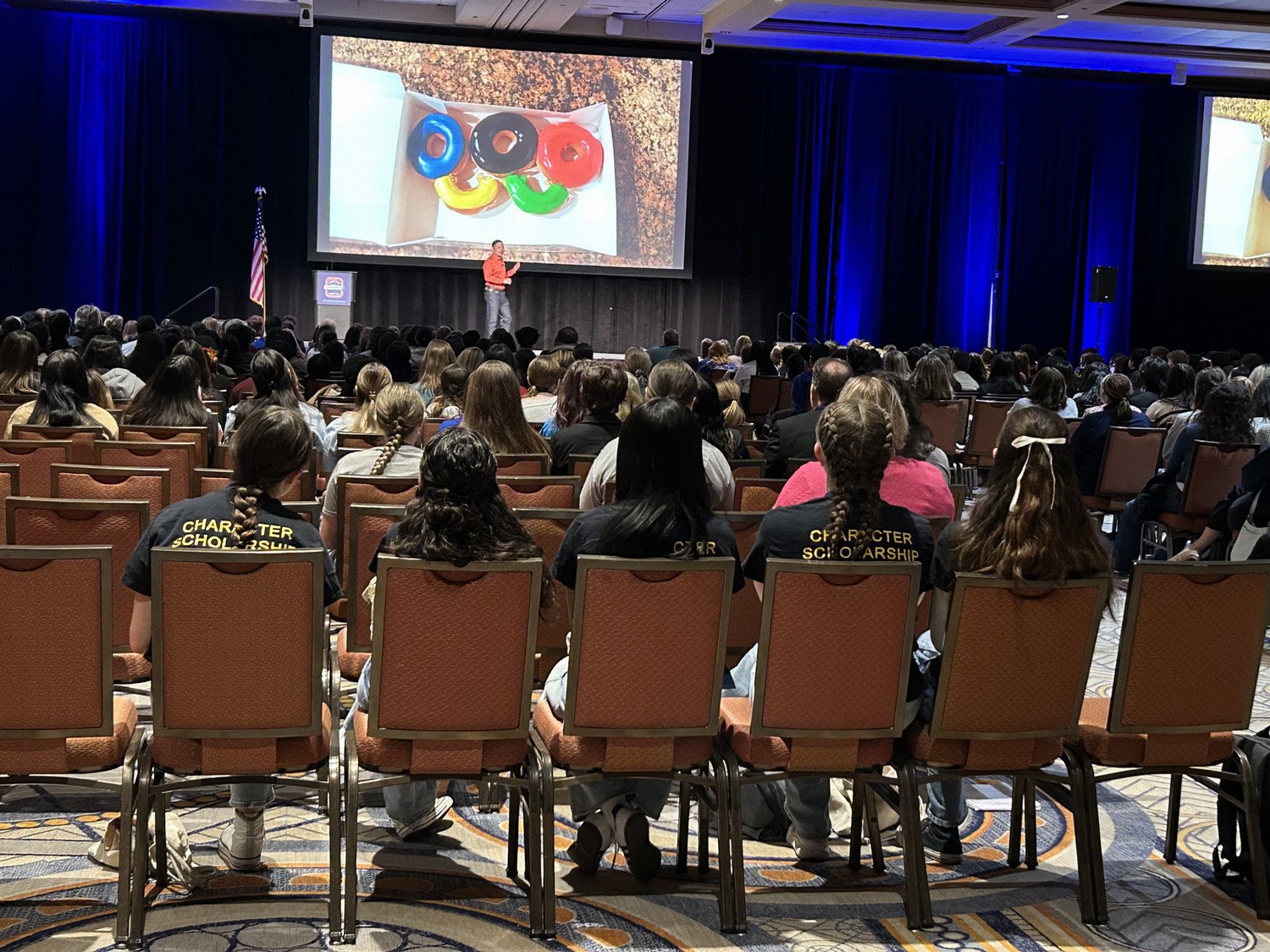 A speaker presents to a large audience seated in a conference hall, with a colorful visual displayed on a screen.