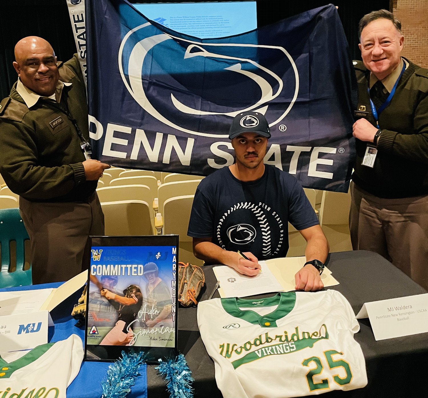 A student athlete signs a commitment letter for Penn State, surrounded by proud coaches and displaying the school flag, while wearing a team jersey.
