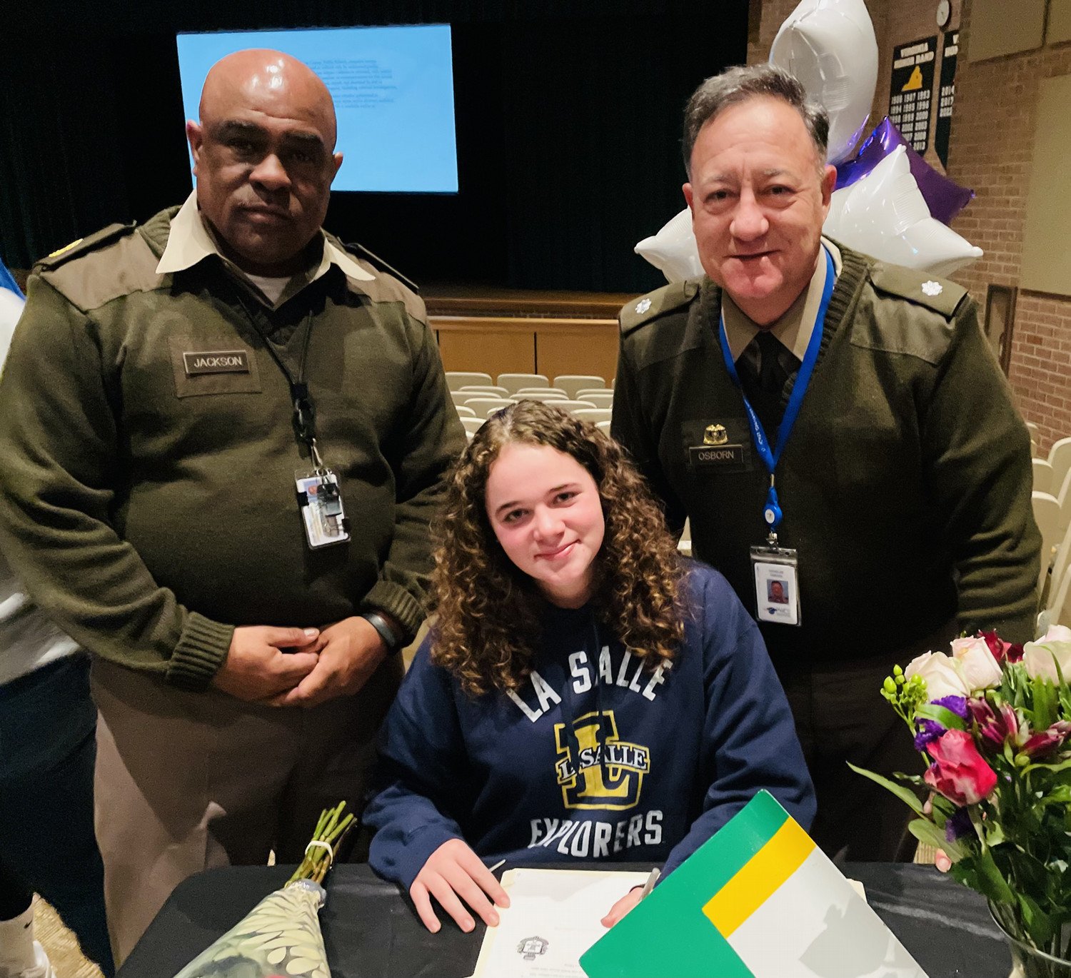 Two uniformed officers standing beside a young woman in a La Salle sweatshirt, who is signing a document at a table adorned with flowers.