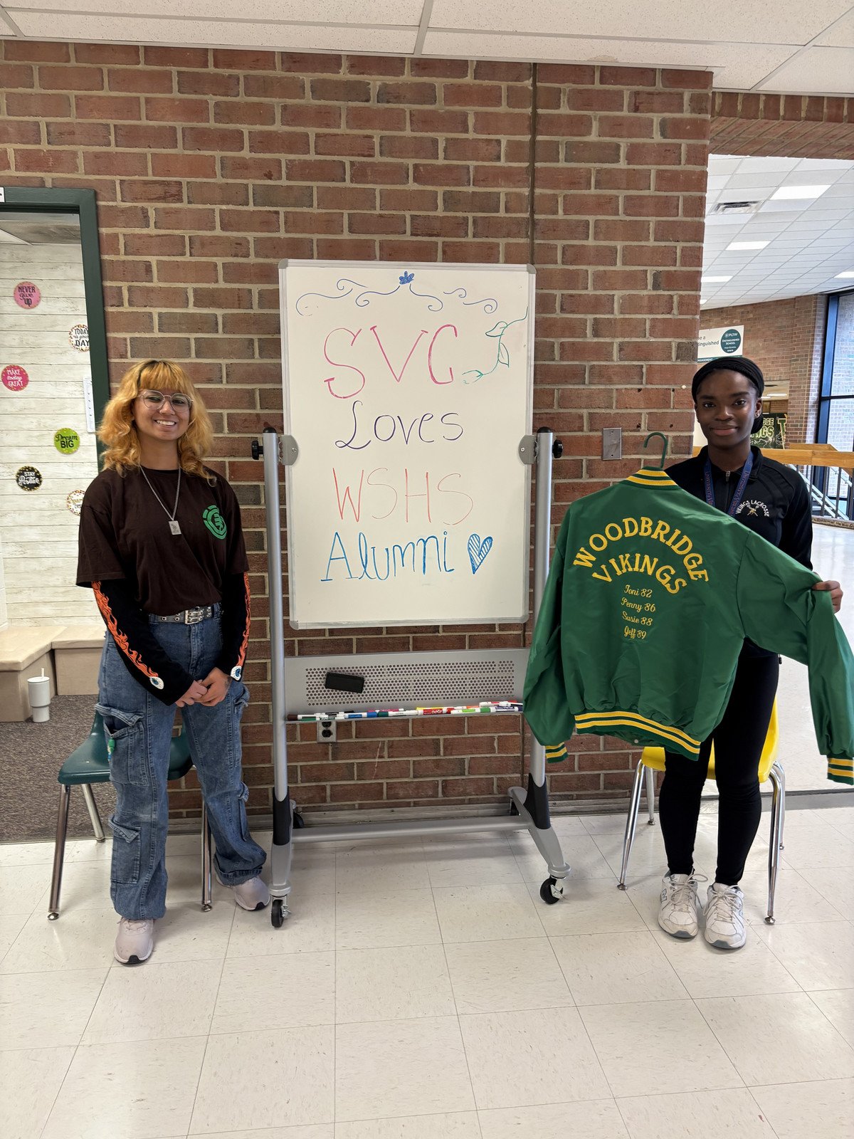 SVC students stand beside a sign that reads "SVC loves WSHS Alumni," holding a green varsity jacket with "Woodbridge Vikings" embroidered on it.