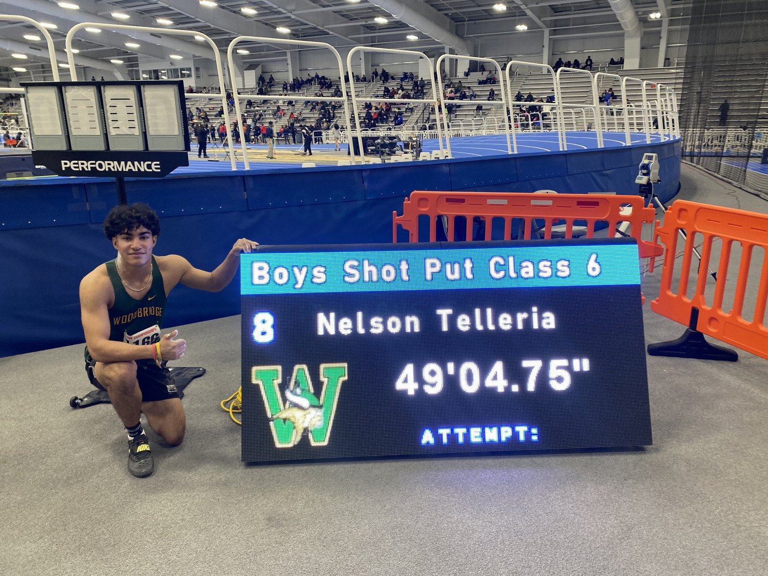 Nelson Telleria kneeling next to a scoreboard displaying his shot put result of 49'04.75" at a Class 6 track event.
