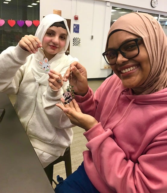 Two young women wearing hijabs proudly hold up their creative keychain crafts. One woman is displaying a colorful bunny design while the other shows a playful panda keychain.
