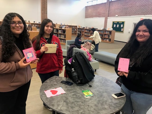 Teenage girls engaging in a creative activity at a library, holding handmade cards and smiling around a table.