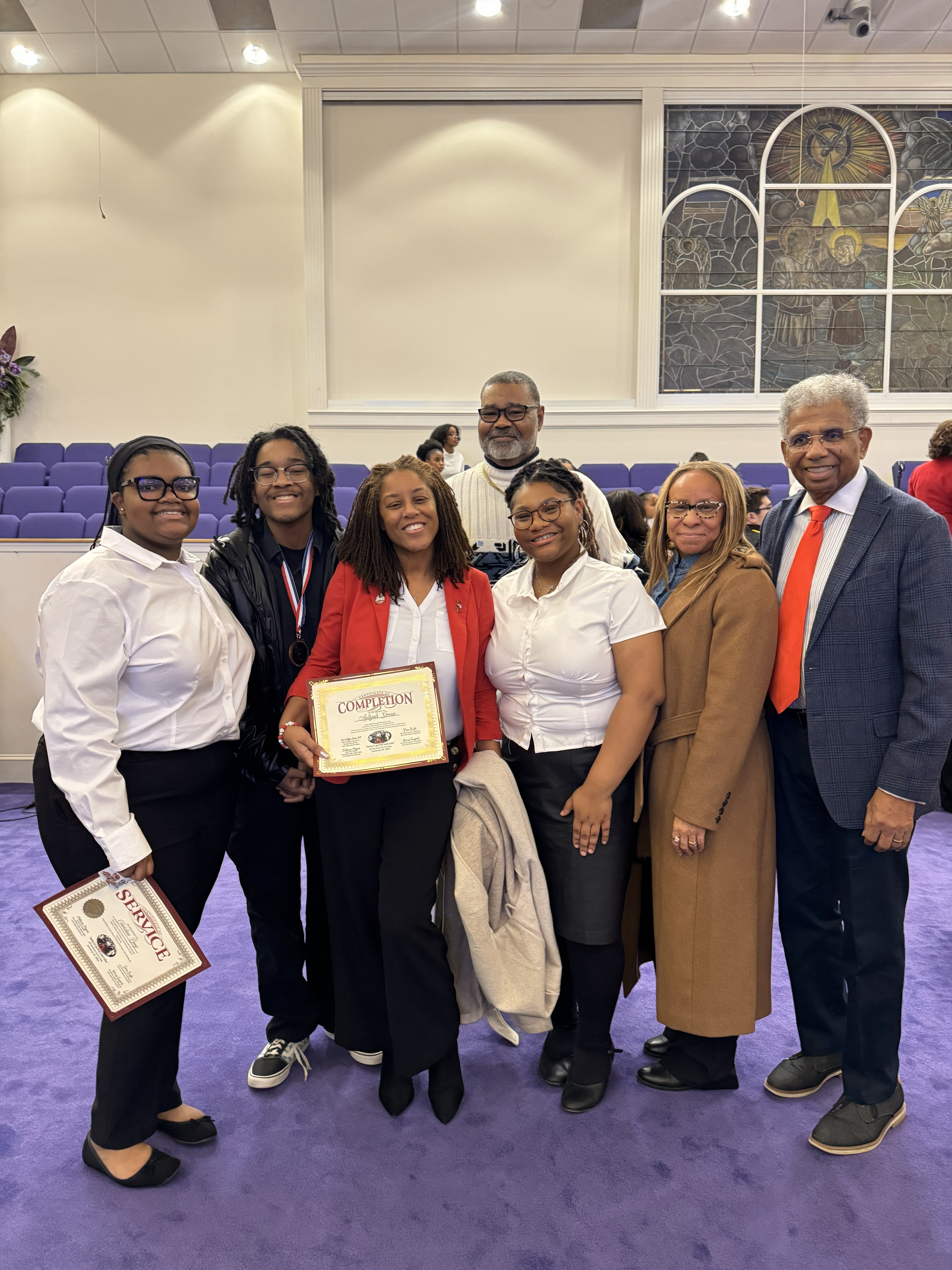 A group of six people celebrating a competition achievement, with one holding a certificate in a church setting.