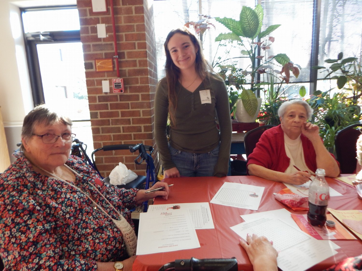 A young woman stands with two elderly ladies at a table, engaging in a community activity surrounded by plants.