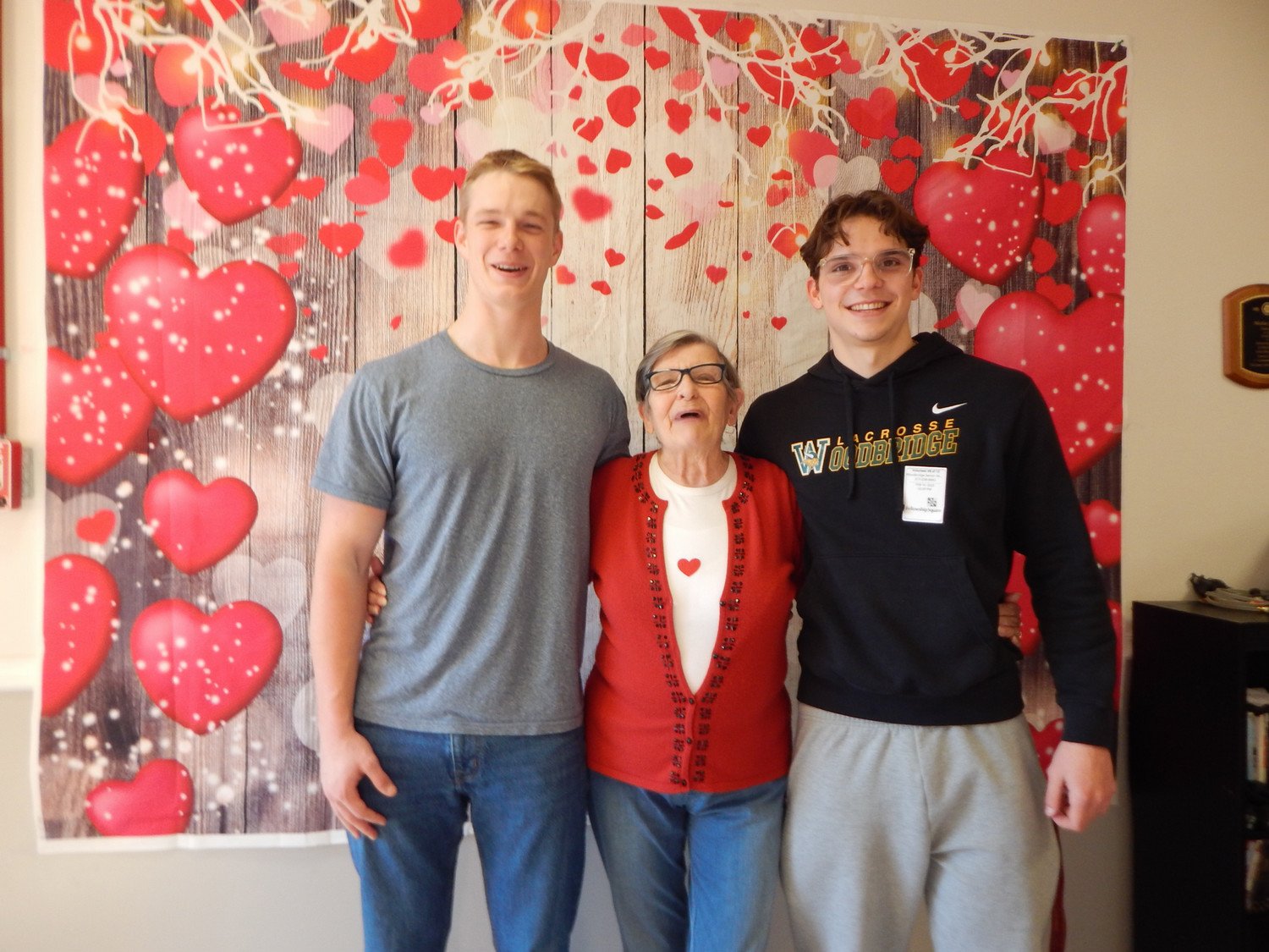 A joyful older woman poses with two young men in front of a backdrop decorated with hearts.