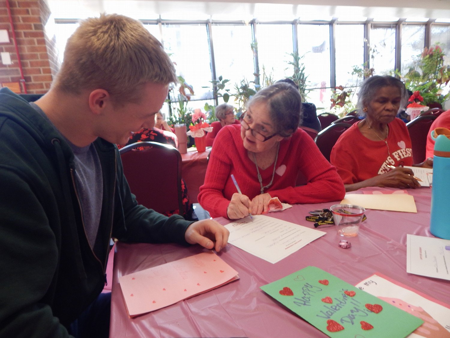 A young man and an elderly woman engage in a heartfelt activity together, with the woman writing on a card decorated with hearts.