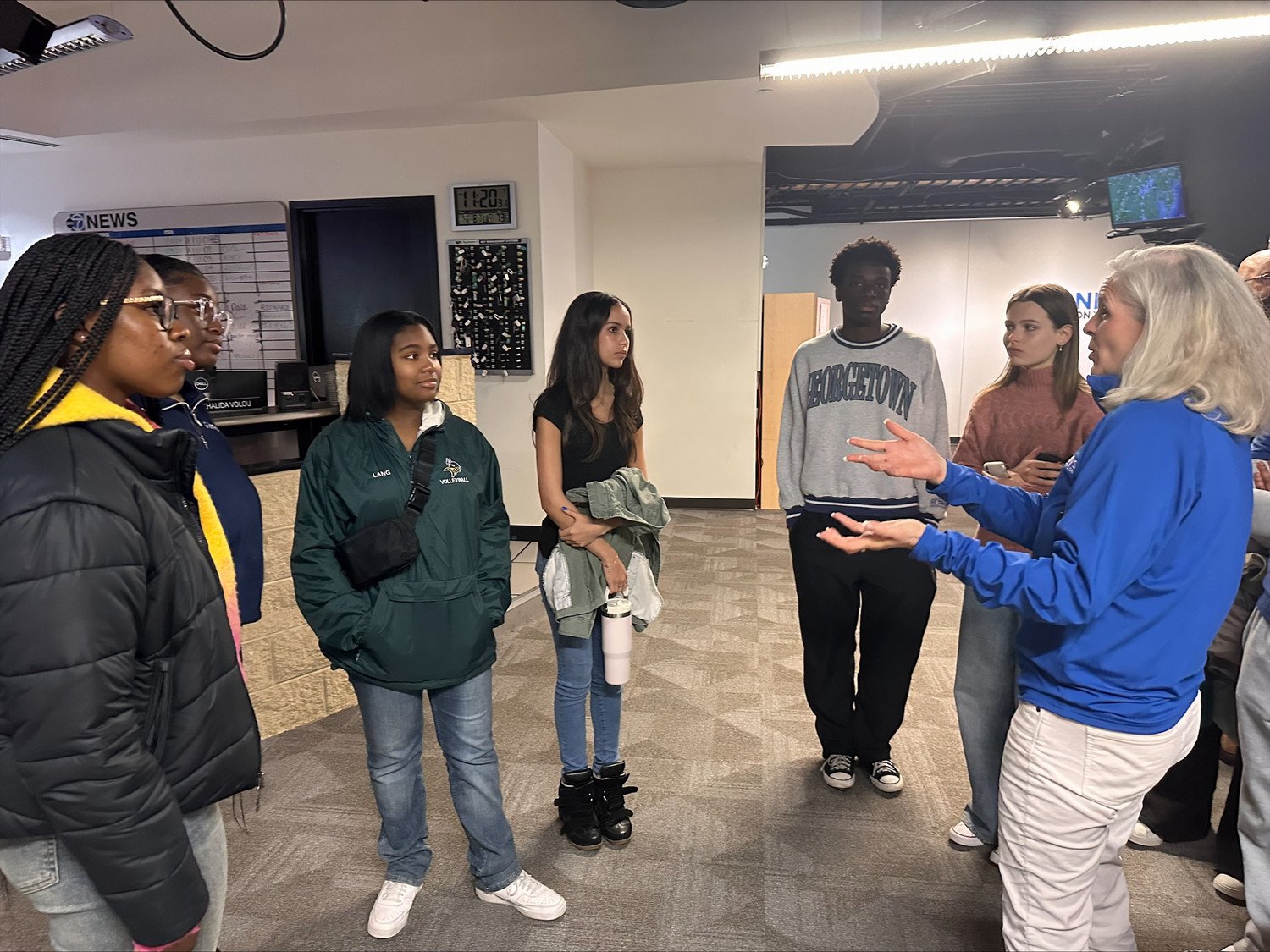 A group of diverse students attentively listens to a woman with long gray hair, who is explaining something in a television studio setting.
