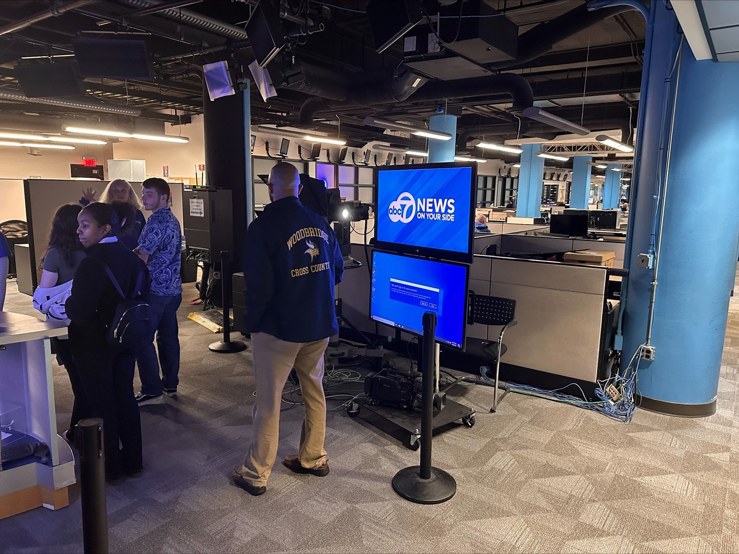 A news studio setup featuring a large screen displaying the ABC 7 News logo, with staff members engaged in a discussion nearby.