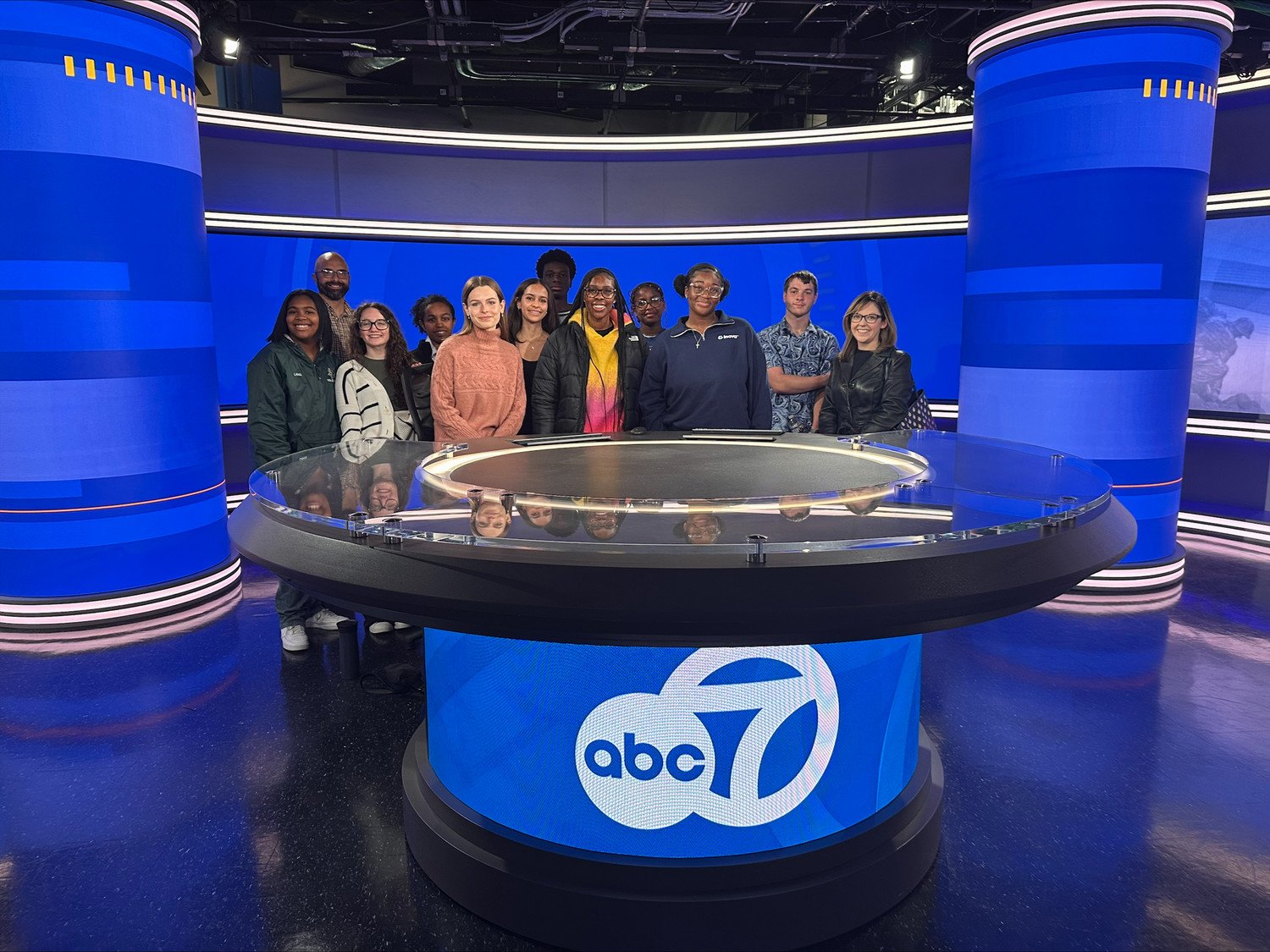 A group of students posing at the ABC 7 news studio, standing around a round glass news desk with blue and white accents.